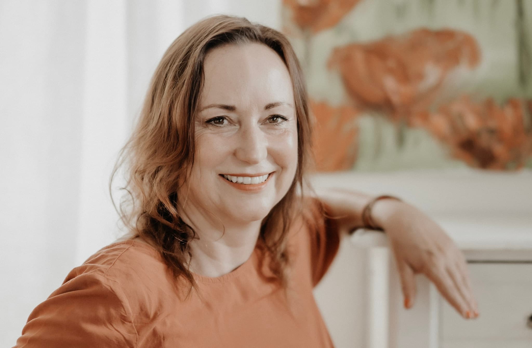 Anja Wolfram lächelnd an einer Kommode, im Hintergrund ein Bild mit roten Blüten