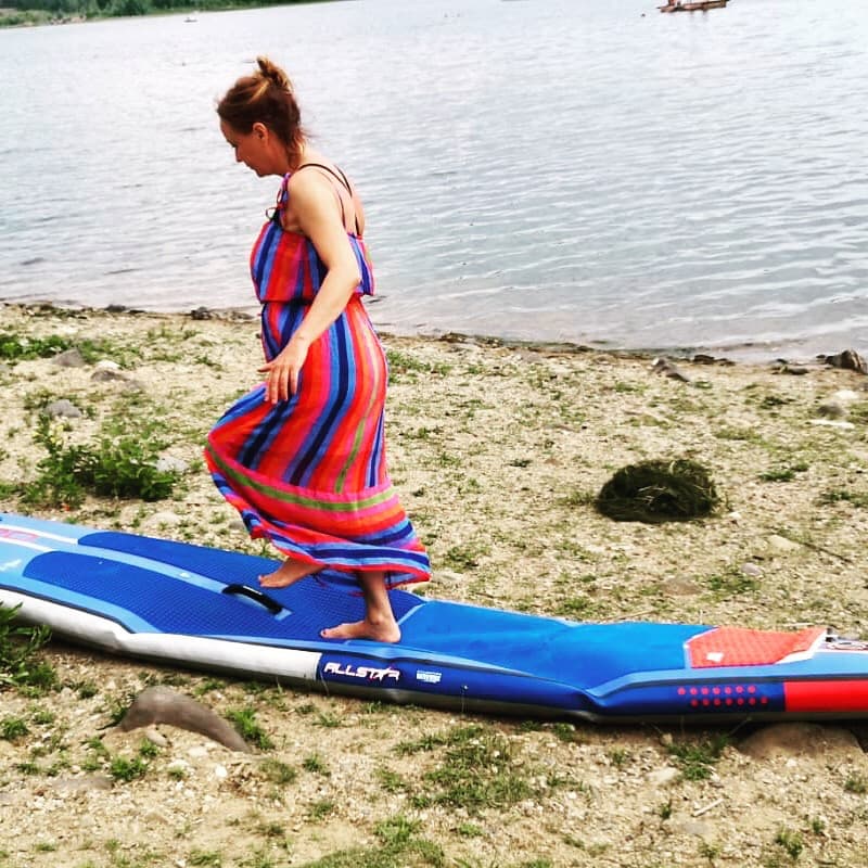 Eine Frau im langen gestreiften Kleid steigt am Seeufer auf ein Stand-up-Paddle-Board