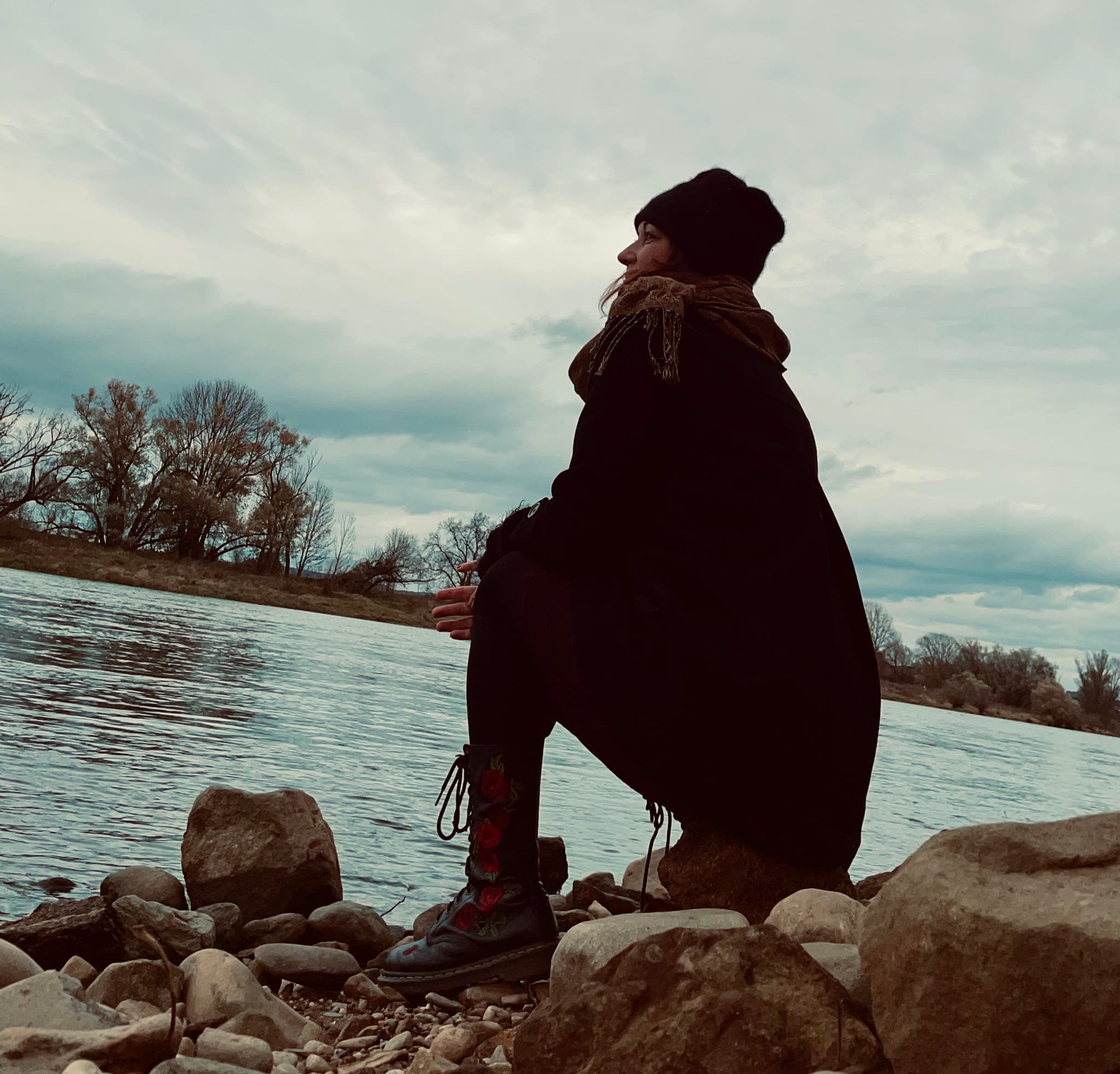 Eine Frau mit Mütze und Mantel sitzt auf Steinen am Elbufer und blickt aufs Wasser