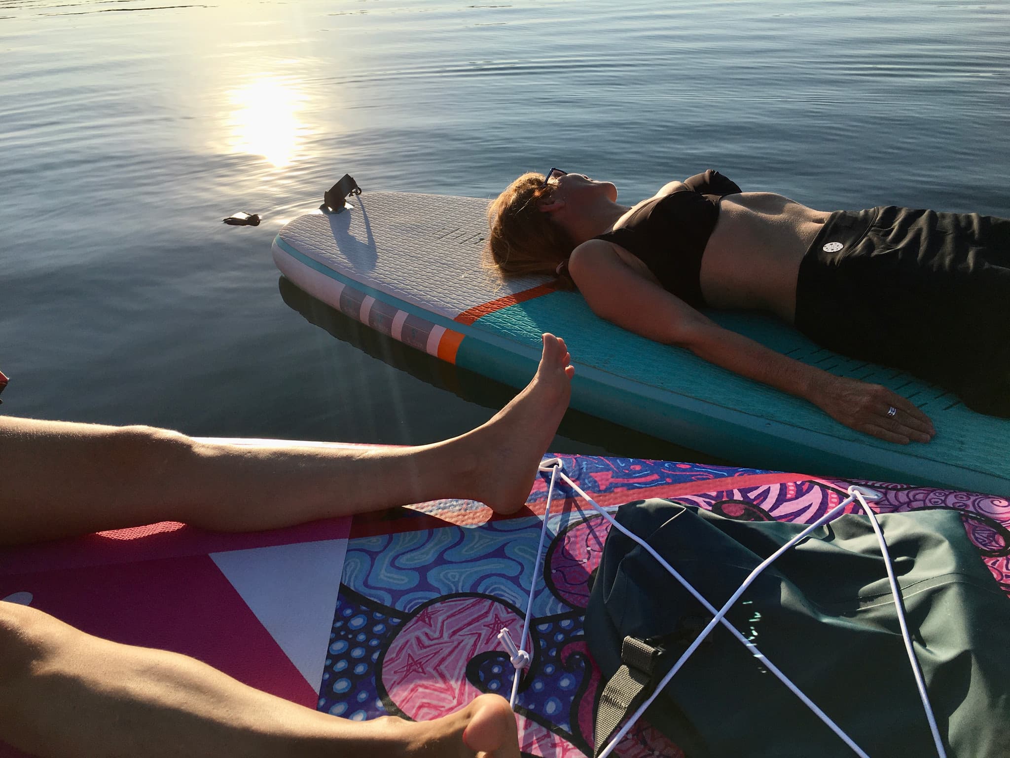 Eine Frau liegt entspannt auf einem Stand-up-Paddle-Board auf dem See, im Gegenlicht der Abendsonne