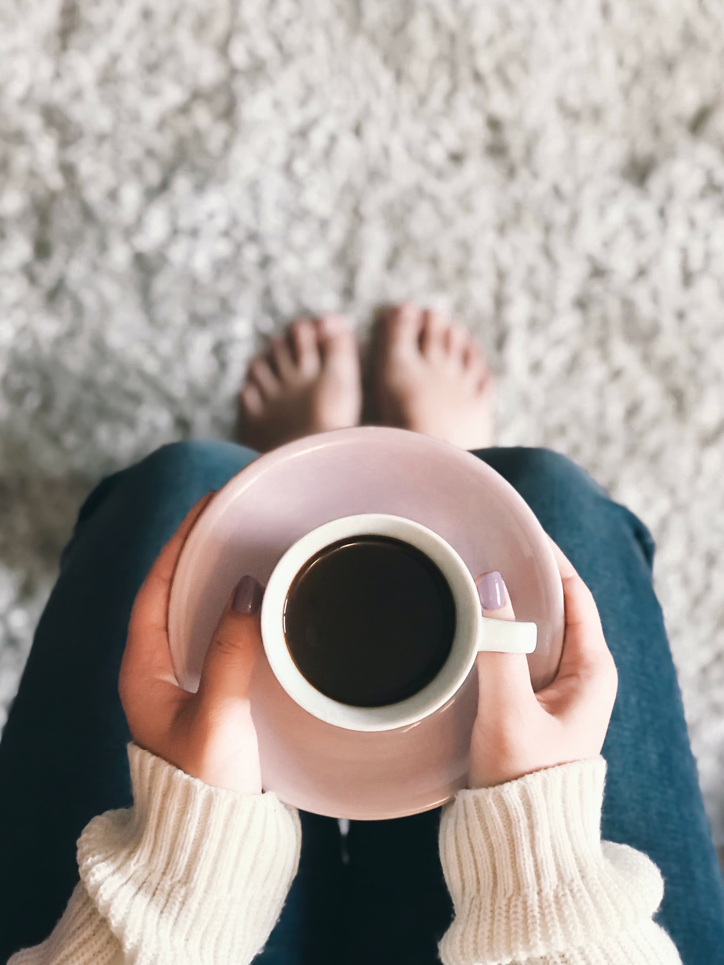 Hände halten eine Tasse Kaffee, von oben fotografiert, darunter nackte Füße