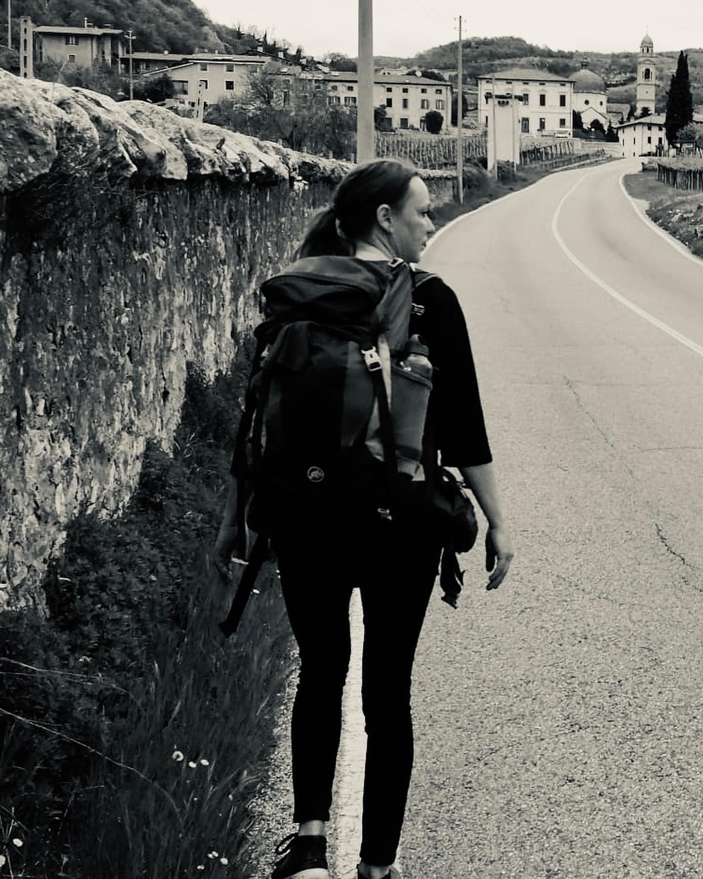 Schwarz-Weiß-Foto: eine Frau mit Rucksack geht eine Landstraße an einer Mauer entlang, im Hintergrund ein italienisches Dorf