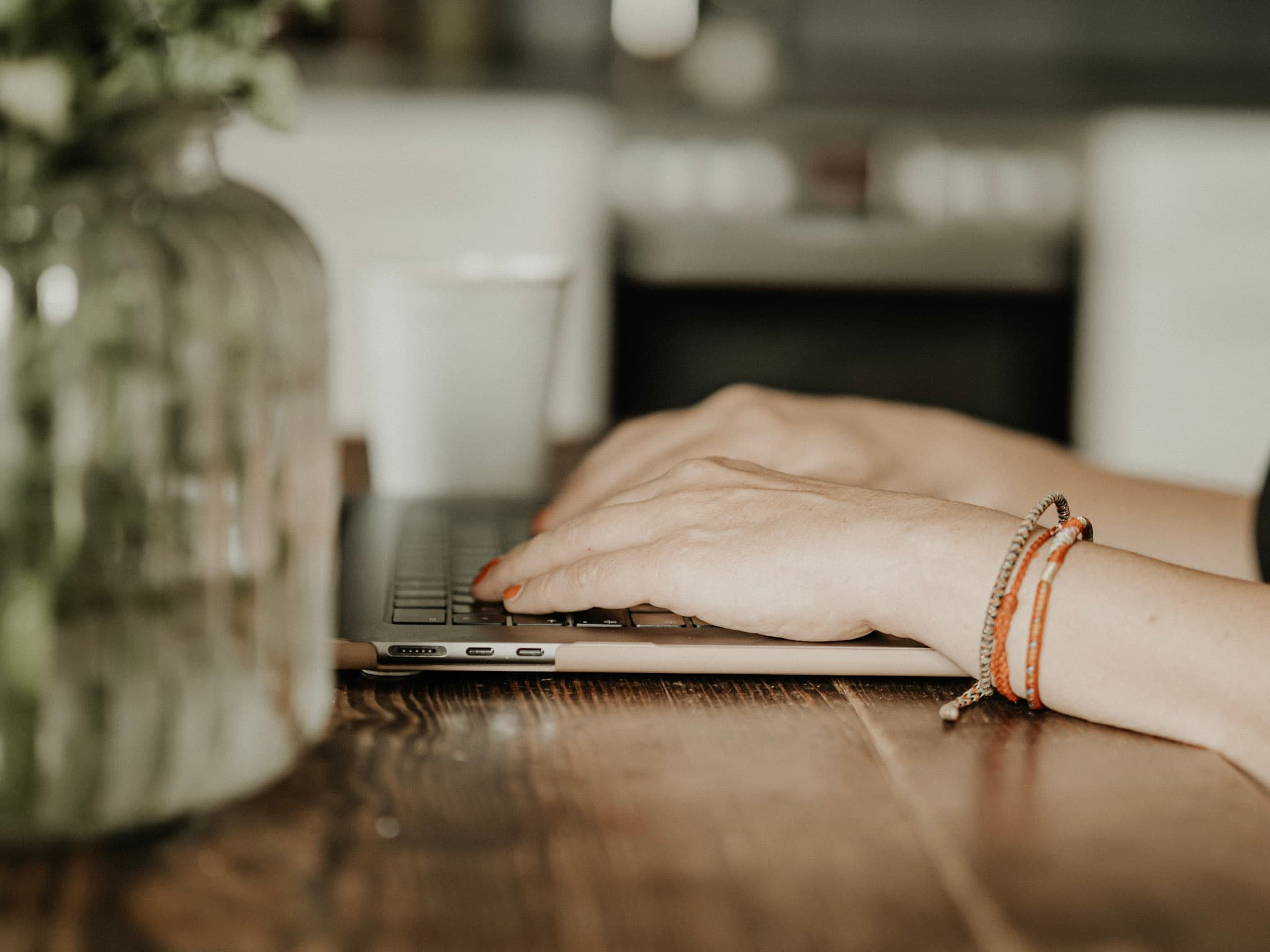 Hand am Laptop auf einem Holztisch, daneben eine Pflanze