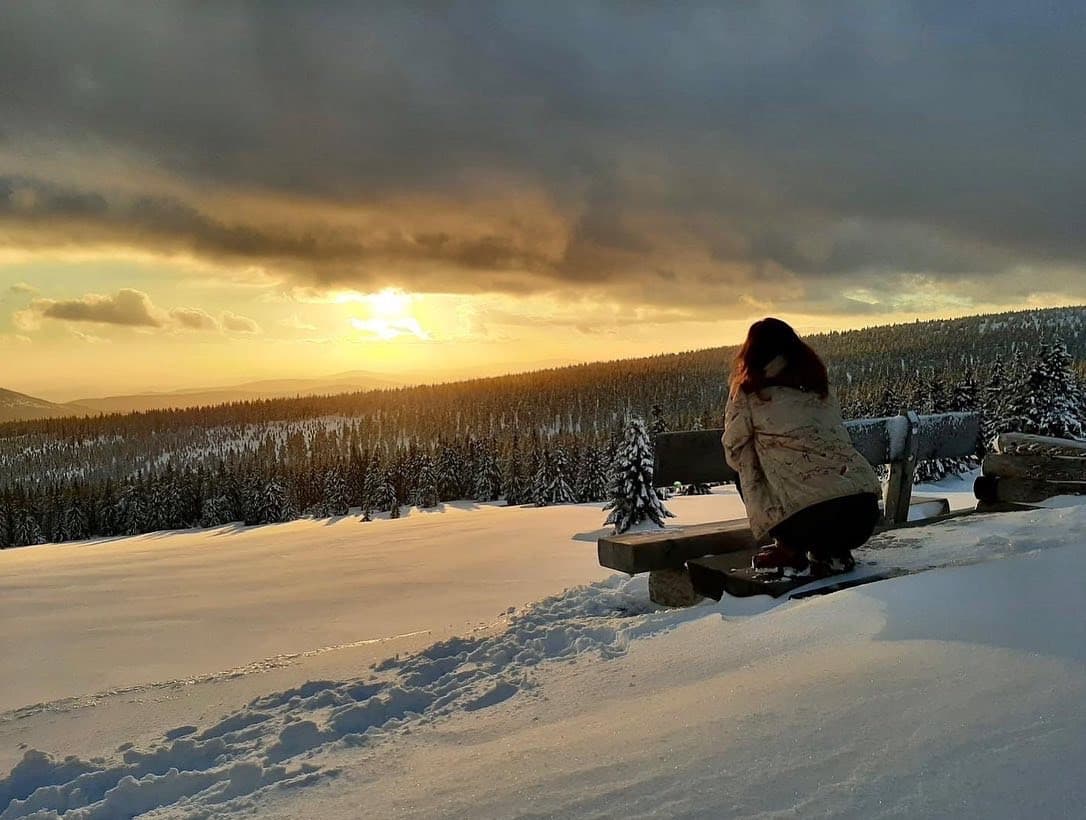 Eine Frau sitzt auf einer Bank im Schnee auf dem Kamm des Riesengebirges, die Sonne geht hinter Wolken unter